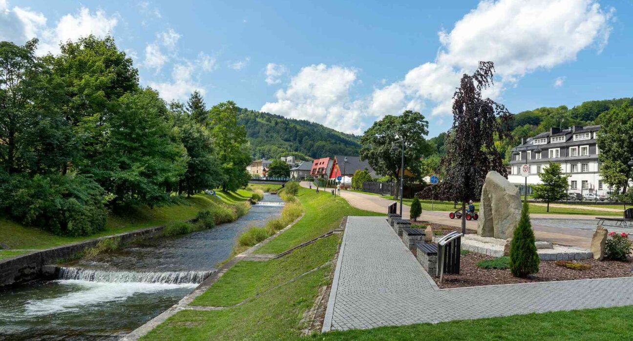 Szczyrk – zagospodarowany bulwar wzdłuż potoku, ścieżka spacerowa i zabudowa apartamentowa na tle gór Beskidu Śląskiego