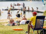 Ratownik w żółtej koszulce z napisem RATO obserwuje kąpielisko pełne plażowiczów i pływaków w słoneczny letni dzień w Poznaniu. W tle jezioro, deska windsurfingowa i pomost.