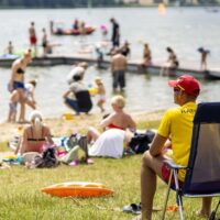 Ratownik w żółtej koszulce z napisem RATO obserwuje kąpielisko pełne plażowiczów i pływaków w słoneczny letni dzień w Poznaniu. W tle jezioro, deska windsurfingowa i pomost.