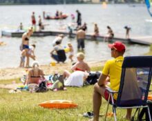 Ratownik w żółtej koszulce z napisem RATO obserwuje kąpielisko pełne plażowiczów i pływaków w słoneczny letni dzień w Poznaniu. W tle jezioro, deska windsurfingowa i pomost.