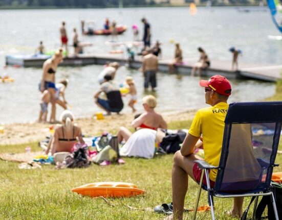 Ratownik w żółtej koszulce z napisem RATO obserwuje kąpielisko pełne plażowiczów i pływaków w słoneczny letni dzień w Poznaniu. W tle jezioro, deska windsurfingowa i pomost.