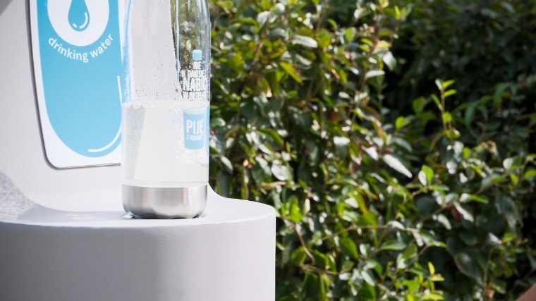Zdrojownik do napełniania butelek z oznaczeniem „drinking water" i logo Aquanet — miejski punkt z darmową wodą pitną w Poznaniu, ustawiony na białym postumencie wśród zieleni.